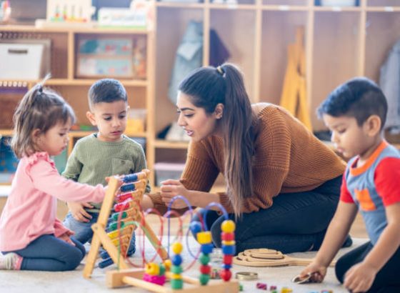 A female Kindergarten teacher of Middle Eastern decent, sits on the floor with students as they play with various toys and engage in different activities.  They are each dressed casually as they learn through their play.
