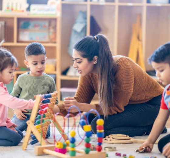 A female Kindergarten teacher of Middle Eastern decent, sits on the floor with students as they play with various toys and engage in different activities.  They are each dressed casually as they learn through their play.
